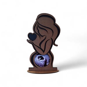 Puede incluir: Silueta de madera de una persona embarazada sosteniendo un corazón. La silueta es marrón con un contorno negro y una imagen de ultrasonido en el área del vientre. La base también es de madera.