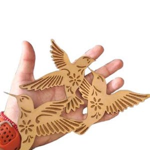 Puede incluir: Tres figuras de colibríes de madera de color marrón claro, planas y cortadas con láser. Cada ave presenta detalles intrincados en las plumas de las alas y un pequeño recorte de flor estilizada en el pecho. Las aves aparecen en posición de vuelo.
