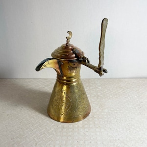 May include: A brass coffee pot with intricate engraved designs. The pot has a long, curved spout and a handle with a decorative finial. The pot is decorated with a crescent moon design on the lid.