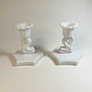 May include: A pair of white candle holders. Each has a dolphin motif supporting a fluted cup for the candle. The holders sit on a hexagonal base. The design is a classic, elegant style, suitable for a dining table.