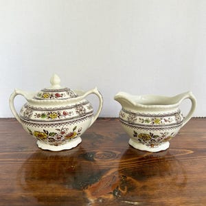 May include: A cream-coloured sugar bowl with a lid and a creamer. Both pieces feature a floral pattern with yellow, red, and green accents. The sugar bowl has two handles and a decorative finial on the lid. The creamer has a spout and a handle.