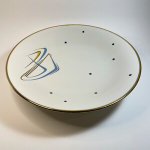 Peut inclure: Une assiette en céramique blanche avec un bord doré et un motif abstrait bleu et jaune. L'assiette est décorée de points noirs.