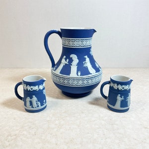 May include: A set of three blue and white ceramic pieces, including a pitcher and two creamer cups. The pitcher has a wide base and a handle, and the cups have a rounded shape with a handle. All three pieces feature a white design of figures and floral motifs on a blue background.