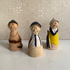 May include: Three quirky ceramic figurines. The first is tan with a black hat and belt. The second wears a blue and white striped shirt and a blue hat. The third has a yellow vest and a white hat. All figures have exaggerated facial features, giving them a comical appearance.