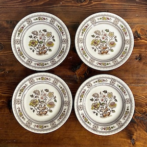 Könnte beinhalten: Vier weiße Keramikteller mit floralem Design. Jeder Teller hat ein zentrales Blumenmotiv in Gelb-, Braun- und Rosatönen, umgeben von einem dekorativen Rand. Die Teller sind auf einer Holzoberfläche angeordnet.