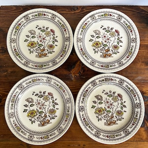 Könnte beinhalten: Vier passende Vintage-Keramikplatten. Jede Platte hat ein zentrales Blumendesign in Gelb, Braun und Rot, umgeben von einem dekorativen Rand. Die Teller stehen auf einer Holzoberfläche.