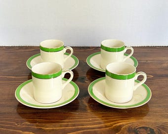 May include: Four cream-colored espresso cups and saucers with green trim. The cups have a cylindrical shape with a handle, and the saucers are round. The green trim is on the rim of the cups and the edge of the saucers. The set is arranged on a wooden surface.