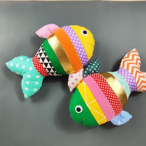 Può includere: Due colorati giocattoli a forma di pesce imbottito. Ogni pesce ha una testa gialla, una fascia dorata e un corpo fatto di vari tessuti fantasia, tra cui pois, triangoli e strisce. Anche le pinne e le code sono decorate.
