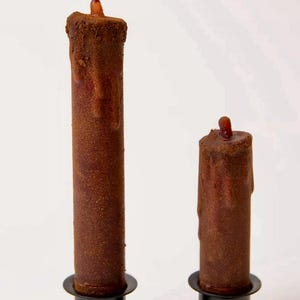 May include: Two brown, textured, cylindrical candles of different heights, each featuring a dark, dripped wax effect and a small, protruding wick at the top. The candles are set into simple, black, circular metal candle holders.