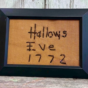 May include: Black framed artwork with the words "Hallows Eve 1772" written in black on a tan fabric background. The frame is rectangular and the artwork is displayed on a wooden surface.