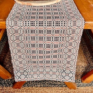 May include: A woven table runner with a geometric pattern in black, red, and cream. The runner is draped over a wooden table, showcasing its intricate design and texture. The runner's dimensions are not visible.