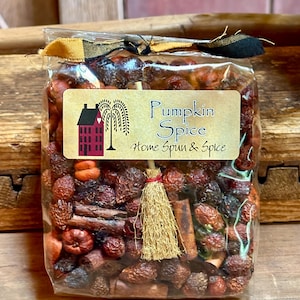 May include: A clear cellophane bag filled with dried spices and decorative elements. The bag is tied with a black and gold plaid ribbon. A label reads "Pumpkin Spice Home Spun & Spice" with a house and tree illustration.