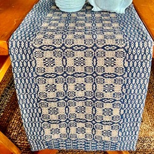 May include: A rectangular table runner featuring a woven pattern in navy blue and beige. The design consists of repeating geometric squares and intricate floral-like motifs arranged in a grid, with a textured border along the sides.