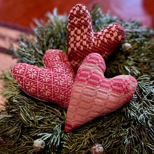May include: Three heart-shaped ornaments with intricate woven patterns in shades of red and cream, resting on a bed of green pine needles. The ornaments have a textured appearance, suggesting a handmade craft.