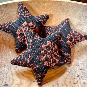 May include: Three star-shaped decorative pillows with a black and orange woven pattern. The pillows are arranged in a wooden bowl, showcasing their textured fabric and geometric design. The stars have a soft, plush appearance.