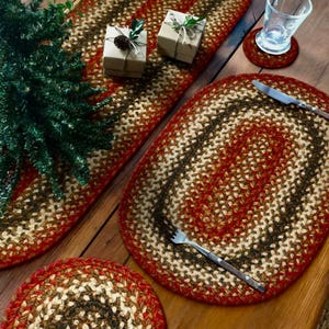 May include: Braided oval placemats and a runner in shades of red, brown, and cream. A matching coaster and a fork and knife are on the table. Two small wrapped gifts with pinecones and greenery are also present.