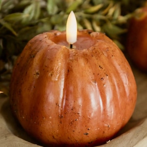 May include: A round, pumpkin-shaped candle with a textured, mottled orange-brown surface and vertical ridges. A small, white, flame-shaped LED bulb sits in the center top, emitting a soft, warm glow, mimicking the appearance of a lit candle.