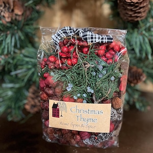 May include: A clear cellophane bag filled with a mix of dried berries, pine needles, and small pine cones. The bag is tied with a black and white checkered ribbon. A label reads "Christmas Thyme, Home Spun & Spice."