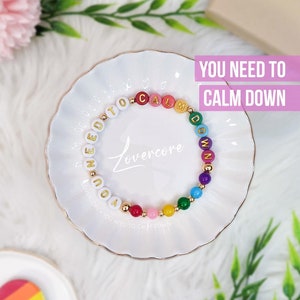 May include: A colorful beaded bracelet with the text "YOU NEED TO CALM DOWN" and "Lovercore" on a white plate. The bracelet features gold accents and rainbow-colored beads. A rainbow heart-shaped object is also visible.