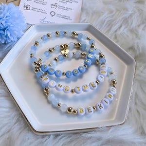 May include: Three beaded bracelets with blue and white beads and gold accents. The bracelets spell out the phrases "Try Try Try", "Find the Joy", and "Be Kind".