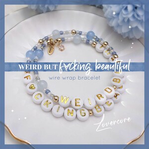 May include: White beaded bracelet with gold lettering spelling "WEIRD BUT F*CKING BEAUTIFUL" on a white background. The bracelet is made with wire and has blue beads.