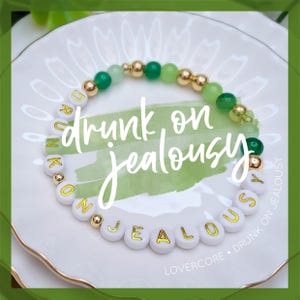 May include: A white beaded bracelet with gold lettering spelling "drunk on jealousy" on green and gold beads. The bracelet is on a white plate with a green background.