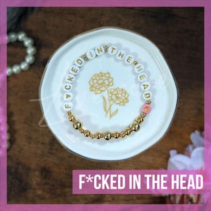 May include: A gold beaded bracelet with white beads that spell out "F*CKED IN THE HEAD" in gold letters. The bracelet is on a white saucer with a floral design.