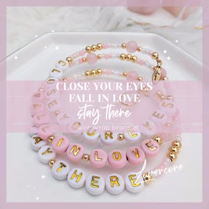 May include: Three layered bracelets with pink and white beads spelling out the phrase "Close your eyes fall in love stay there". The bracelets are made with gold wire and have small pink and white beads.