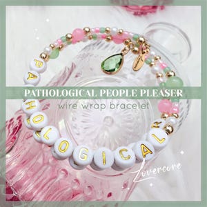 May include: A pink and green wire wrap bracelet with white beads that spell out "Pathological People Pleaser" in gold lettering.