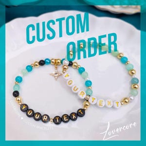 May include: Two custom beaded bracelets with the text "YOUR TEXT". One bracelet has black beads with gold lettering, the other has white beads with gold lettering. Both bracelets have gold and turquoise beads. The words "CUSTOM ORDER" are in the image.