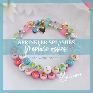 May include: A colorful wire wrap bracelet with the words "SPRINKLER SPLASHES fireplace ashes" spelled out in gold letters on pastel-colored beads. The bracelet also features a small blue teardrop charm and a gold heart charm.