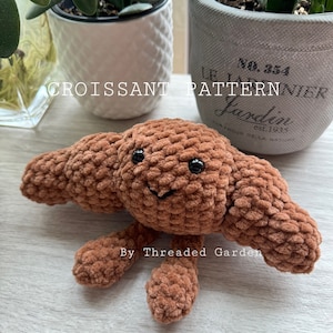 May include: A brown crocheted croissant plush toy with a smiling face. The toy has two small arms and two legs. The toy is on a light brown wooden surface. The text "By Threaded Garden" is visible below the toy.