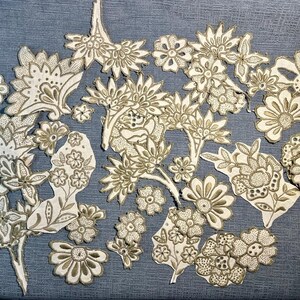 May include: A collection of intricate, beige-colored paper cutouts featuring floral and paisley designs. The cutouts are arranged on a blue fabric background.