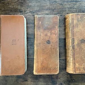 May include: Three antique books with worn, brown leather covers are arranged on a wooden surface. The books have embossed designs and handwritten text. The covers show signs of age and use, with visible wear and tear.