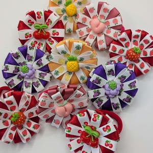 Puede incluir: Una colección de nueve coleteros circulares con cintas superpuestas con estampados de frutas como cerezas, fresas, uvas, melocotones y piñas, cada uno centrado con un dije de plástico tridimensional con forma de fruta a juego.