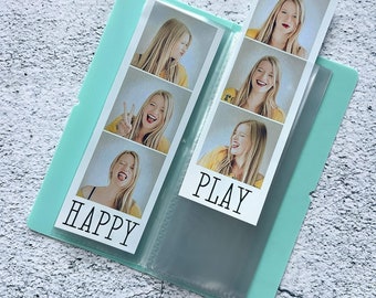 Puede incluir: Un álbum de fotos turquesa con fundas de plástico transparente para guardar fotos. El álbum tiene las palabras "HAPPY" y "PLAY" impresas en la portada.
