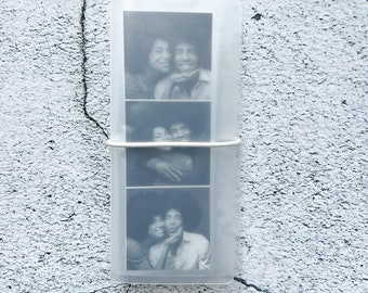 Puede incluir: Un portafotos de plástico transparente con tres tiras de fotos en blanco y negro de una pareja. Las fotos muestran a la pareja en diferentes poses, incluyendo un primer plano de sus caras y una foto de ellos abrazándose.