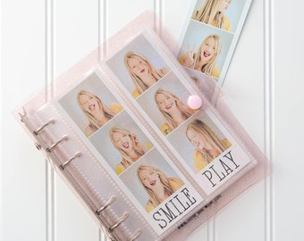 Puede incluir: Un álbum de fotos rosa con purpurina, con una cubierta de plástico transparente y anillos plateados. El álbum tiene una pegatina blanca en la parte delantera que dice "Smile Play" con una dirección de sitio web debajo. El álbum está lleno de tiras de fotos de una mujer haciendo diferentes expresiones faciales.