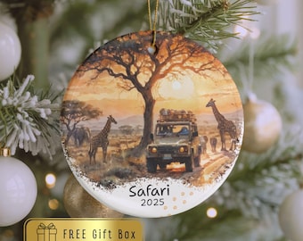 May include: Round ceramic Christmas ornament featuring a safari scene with giraffes, an elephant, and a vehicle. The ornament has the word "Safari" and the year "2025" printed on it. The background shows a sunset with a large tree.