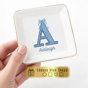 May include: A square white ceramic trinket dish with a gold-toned rim. The center features a large blue striped letter A topped with a blue bow graphic and the name Ashleigh printed below. A gold rectangular label displays Choose Dish Shape.