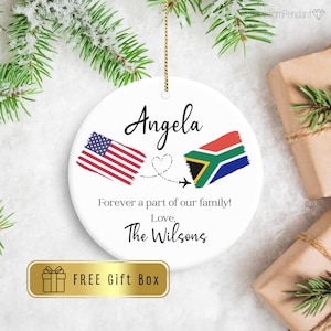 May include: A white ceramic ornament with the name "Angela" in black script, featuring the American and South African flags. The text "Forever a part of our family! Love, The Wilsons" is also present. A gold box with the text "FREE Gift Box" is at the bottom.