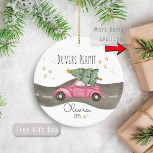 May include: A round ceramic Christmas ornament with a pink car carrying a Christmas tree. The ornament reads "Drivers Permit" and includes the name "Olivia" and the year "2025". The ornament has a gold string for hanging.