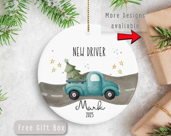 May include: A white ceramic Christmas ornament with a teal pickup truck carrying a Christmas tree. The ornament reads "NEW DRIVER" and "Mark 2025" with gold stars. A gold string is attached for hanging. The words "More Designs available" and "Free Gift Box" are also visible.