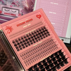 Könnte beinhalten: Ein rosa und transparentes Plastiketui mit Reihen von schwarzen Wimpernextensions. Auf der Vorderseite des Etuis steht der Text "Bahama Lashes Russian Volume Lower Lash". Das Etui ist außerdem mit einem rosa Herz und einer Palmengrafik verziert.
