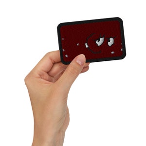 May include: A rectangular, maroon embroidered patch with a black border. The patch features a cartoon face with white eyes and a grumpy expression. The patch is held in a hand.