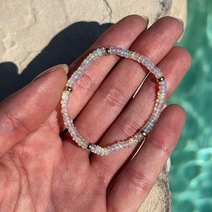 May include: A delicate bracelet featuring small, iridescent opal beads strung together. The bracelet is accented with small gold-colored beads. The jewelry is displayed against a background of a hand and a pool of water.