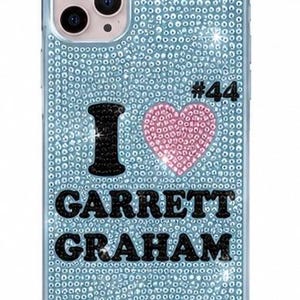 May include: A light blue phone case covered in sparkling rhinestones. The design features a black letter I, a pink rhinestone heart, and the black text GARRETT GRAHAM with the number #44 in black, all set against the light blue crystal background.