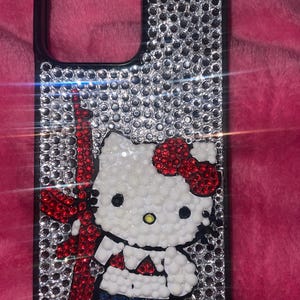 May include: A bedazzled phone case with a black border and a sparkling silver background. The case features a white Hello Kitty character with a red bow and a red and blue weapon. The case is covered in small, shiny rhinestones.