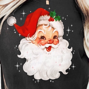 May include: Black t-shirt featuring a vintage Santa Claus graphic. The Santa illustration has a red hat, white beard, and a smiling face. The design includes holly and star accents, with the text "Joy to You" at the bottom.