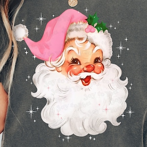 May include: Dark gray t-shirt featuring a vintage Santa Claus graphic. Santa is depicted with a pink hat, rosy cheeks, and a fluffy white beard. The design includes sparkling star accents and the text "Joy to You".